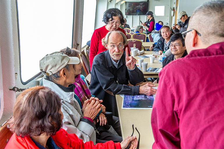 Group of people seated and gathered around a table inside of a ship giving their attention to a man that is speaking.